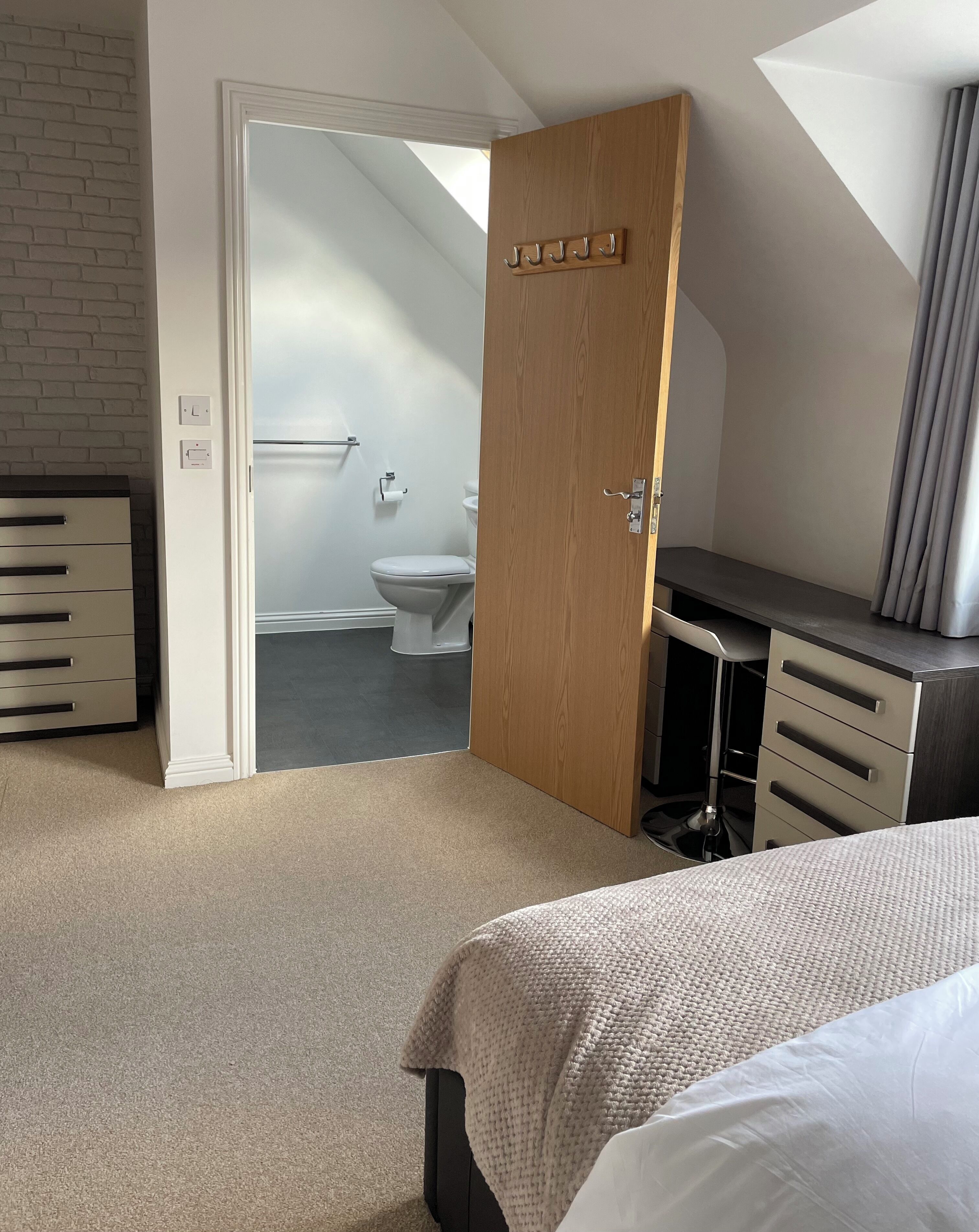 A bedroom with a bed, a wooden door, a desk, and a view into the bathroom.