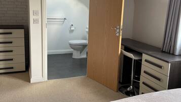 A bedroom with a bed, a wooden door, a desk, and a view into the bathroom.