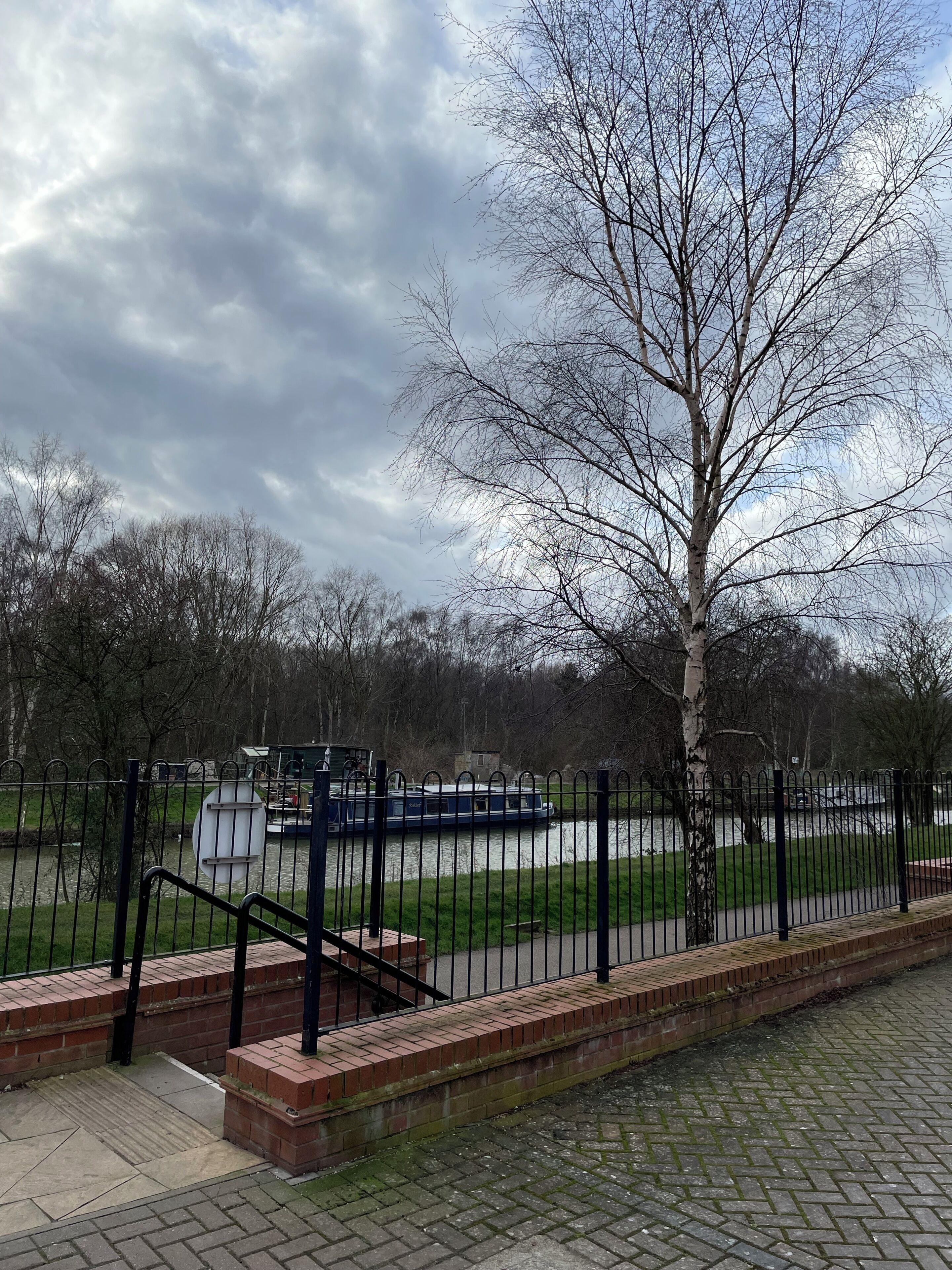 A park with a paved walkway, a metal fence, and a river with a boat.