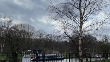 A park with a paved walkway, a metal fence, and a river with a boat.