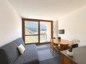 Ett modernt rum med en grå soffa, ett matbord i trä med stolar, en liten krukväxt och en tv som är monterad på väggen.