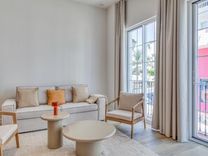 A modern living room with a sofa, armchair, and coffee table, large windows with curtains, and a view of the outside. - Spacious studio near the port - Estepona (Estepona)