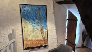 Une pièce avec un mur blanc à texture, un plancher en bois, un fauteuil gris, une petite table blanche et un tableau représentant un ciel bleu et des arbres.