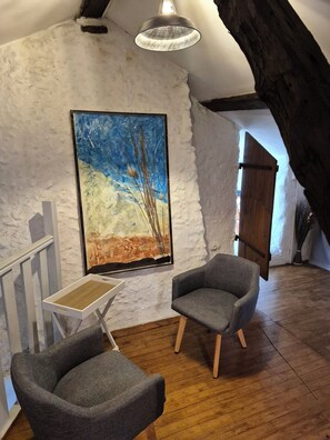 A room with a white textured wall, a wooden floor, a gray armchair, a small white table, and a painting of a blue sky and trees. - GITE IN THE HEART OF THE PERIGORD BLANC FOR UP TO 6 PEOPLE (Neuvic)