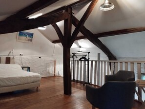 Ein Schlafzimmer im Loft-Stil mit Holzbalkendecke, einem Bett, einem Sessel, einer Pendelleuchte und Blick ins Freie.