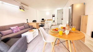 Een moderne woonkamer met een bank, een eettafel met stoelen, een keukenhoek en een slaapkamer met een bed en nachtkastje.