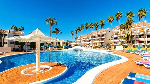 Zona de piscina en un complejo turístico, con una piscina circular, sillas de descanso y palmeras.