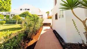 Un sendero conduce a un edificio de fachada blanca, rodeado de vegetación y una valla.