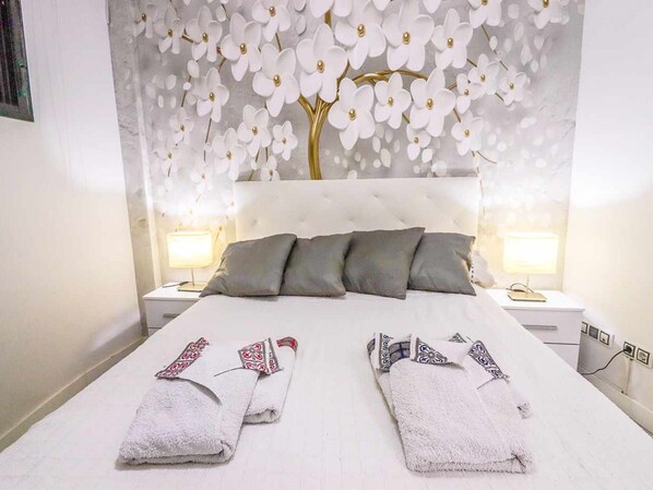 A hotel room with a white bed, grey pillows, two bedside tables with lamps, and a wall with a floral design. - Beautifully decorated apartment in Adeje, close to beach (Adeje)
