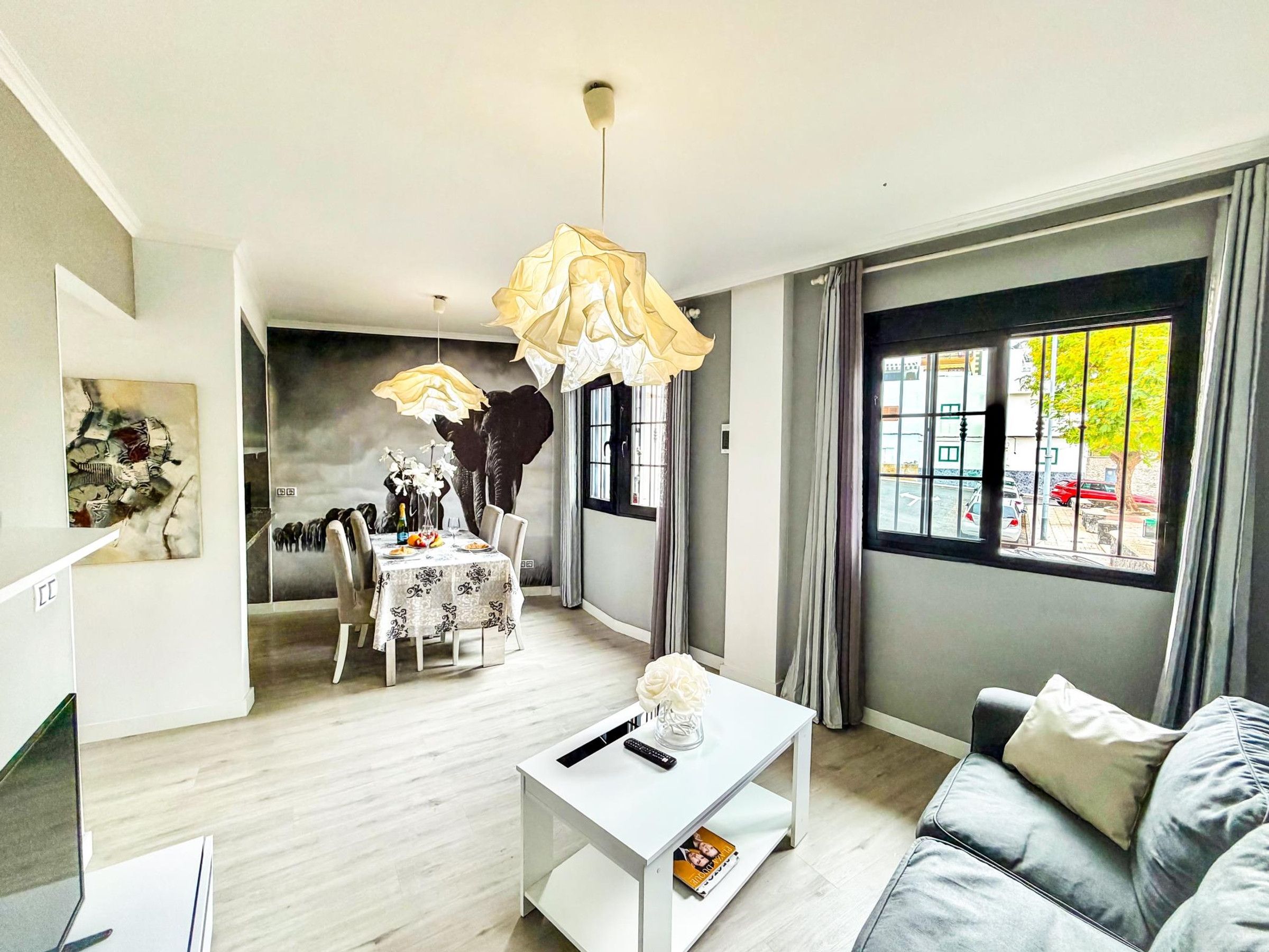 A modern living room with a dining area, a sofa, a coffee table, and a view through a window.