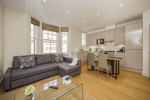 Een moderne woonkamer met een grijze bank, een glazen salontafel en een keukenhoek met witte kasten en een magnetron.