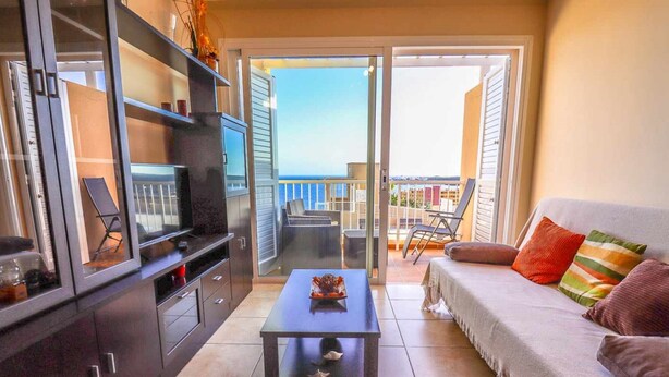 Una sala de estar con un sofá, una mesa de centro y un televisor. Hay una puerta corrediza de vidrio que da a un balcón con mobiliario exterior.