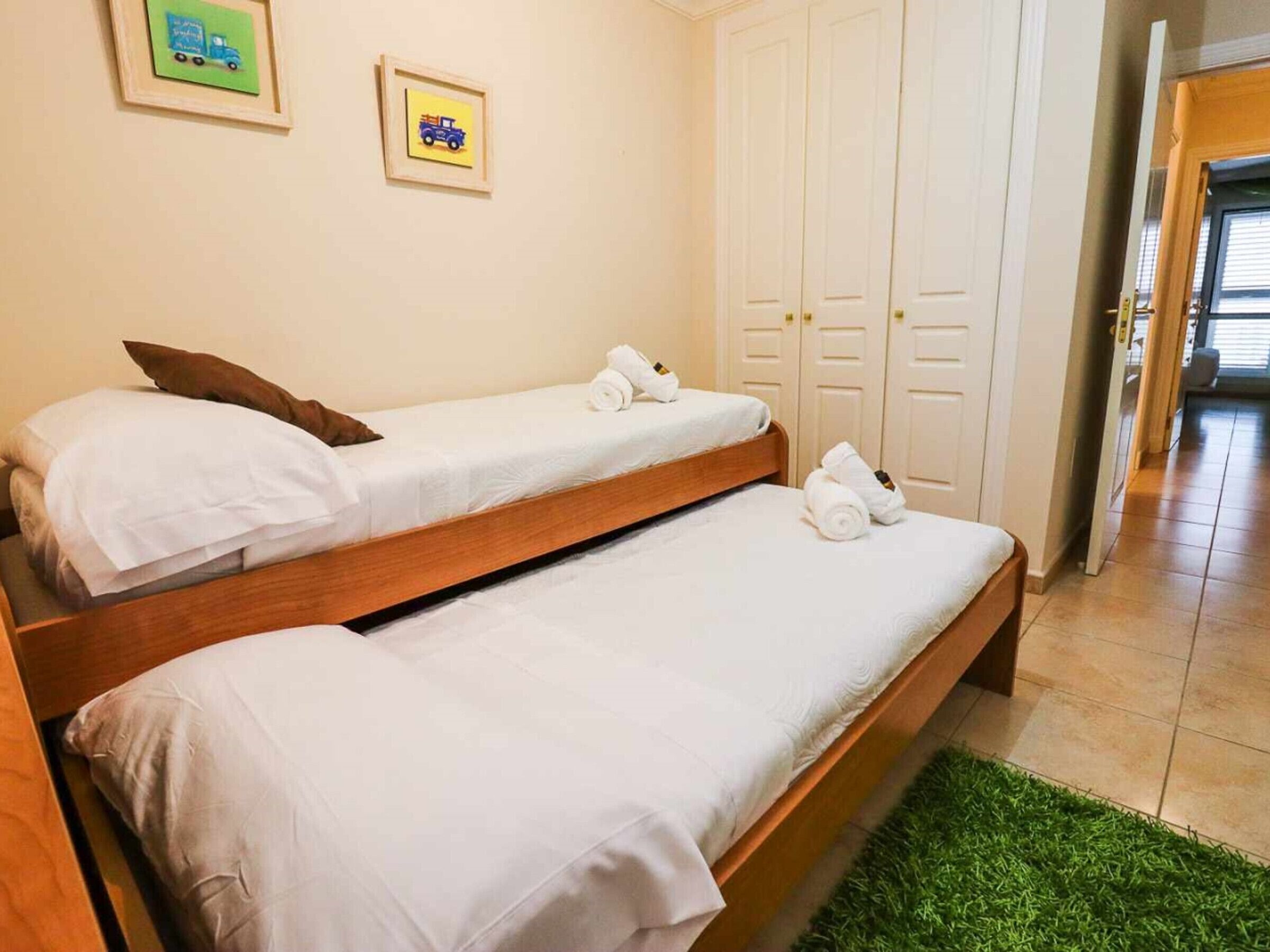 Una habitación con dos camas individuales, ropa de cama blanca, un tapete verde y dos cuadros en la pared.