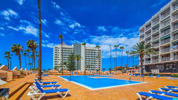 Area piscina di un hotel con lettini, palme e cielo azzurro.