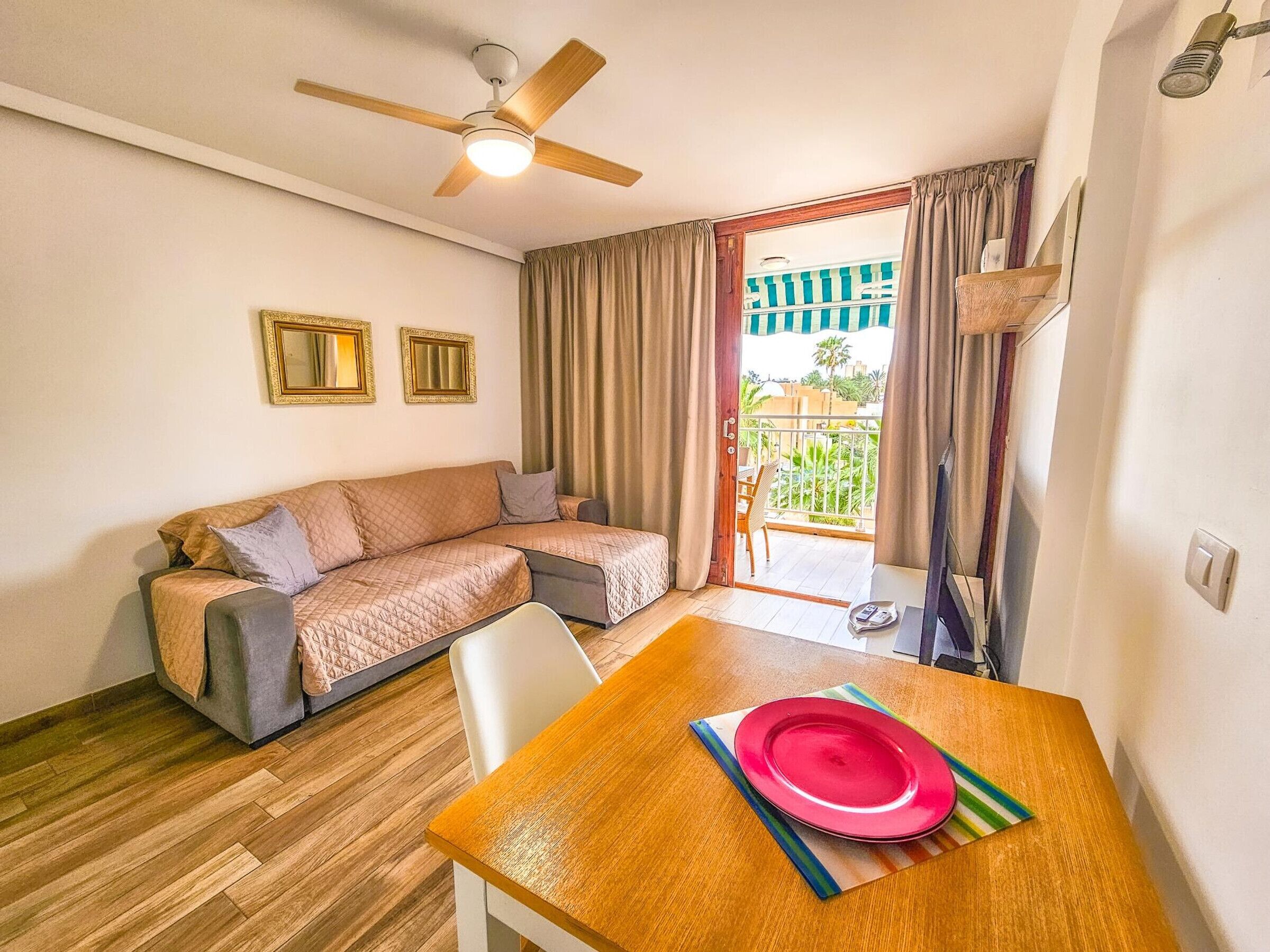 A room with a wooden floor, a sofa, a dining table with a red plate, a ceiling fan, and a view of a balcony with curtains.