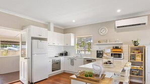A modern kitchen with white cabinets, a refrigerator, oven, microwave, and a coffee maker. There is a window with a view of greenery outside. - The Art Lovers Cottage in Mandurah 1BR (Dudley Park)