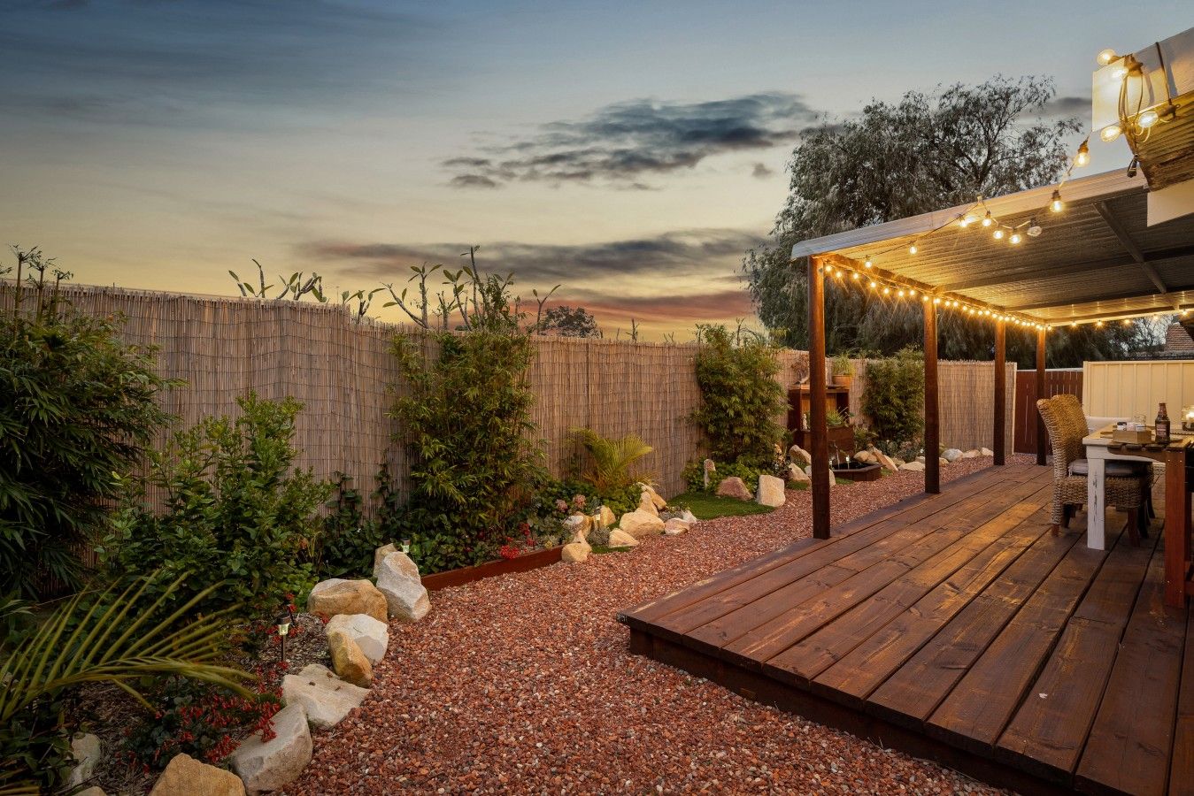 Une terrasse en bois agrémentée de guirlandes lumineuses, un espace salon abrité et un jardin avec des rochers et des plantes.