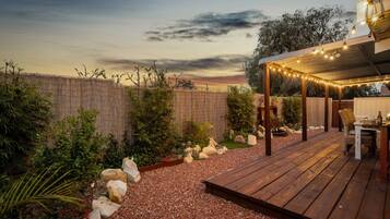 Une terrasse en bois agrémentée de guirlandes lumineuses, un espace salon abrité et un jardin avec des rochers et des plantes.