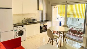 Cozinha compacta com máquina de lavar, micro-ondas e fogão. Possui área de refeições com mesa e cadeiras, e uma varanda com toldo listrado amarelo.