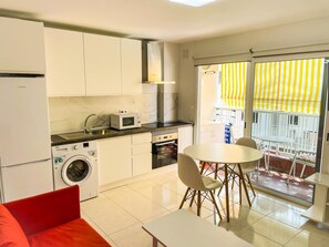 A compact kitchen with a washing machine, microwave, and stove. There is a dining area with a table and chairs, and a balcony with a yellow-striped awning. - Spacious studio 100m from beach, ideal for couples (Los Cristianos)