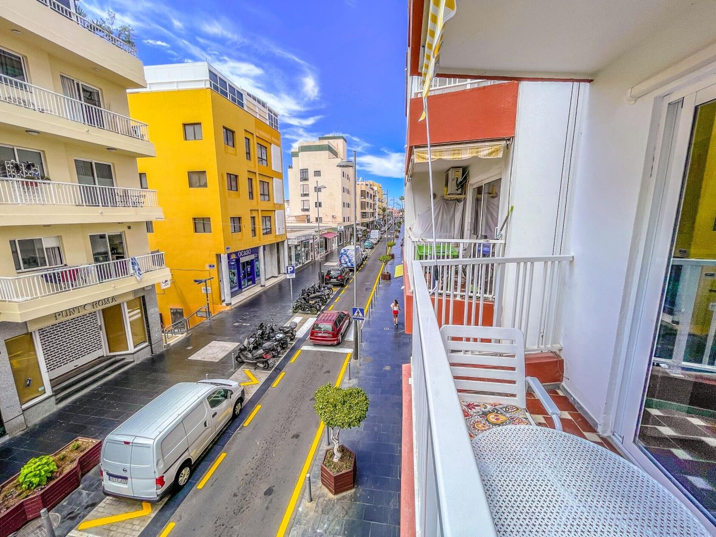 Une rue étroite, bordée de bâtiments des deux côtés, un fourgon blanc garé sur le bord, et un balcon donnant sur la rue.