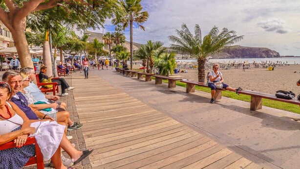 Um calçadão de madeira com bancos, palmeiras e uma praia ao fundo.
