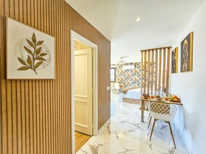 Ein Hotelzimmer mit einer Wand aus Holzpaneelen, einem Bett mit weißer Bettdecke, einem Essbereich mit Tisch und Stühlen sowie einer Wand mit tropischem Wandpapier.