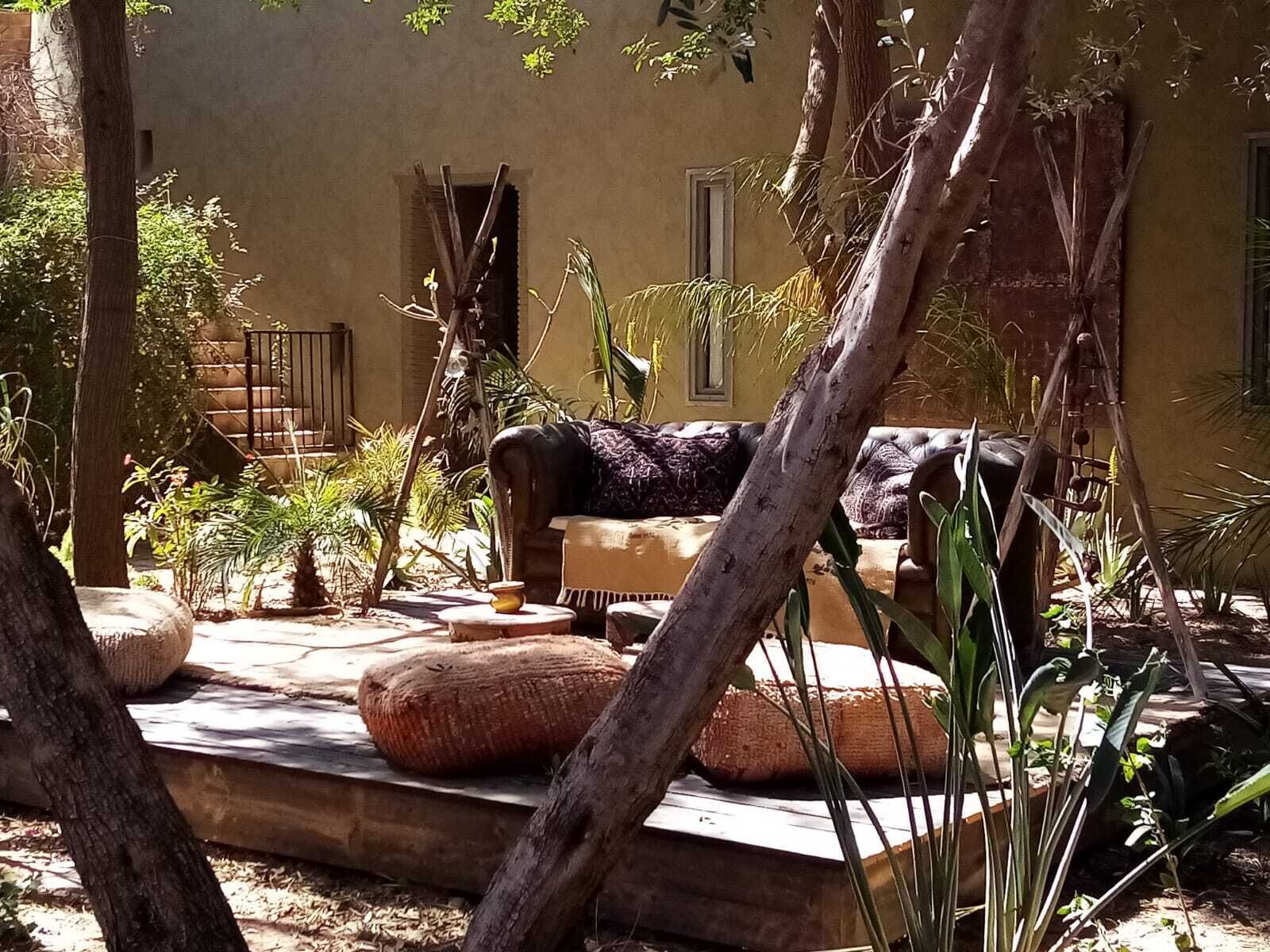 Une cour avec un canapé, une petite table et une chaise. On aperçoit des plantes et un arbre au fond.