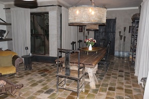 A rustic dining area with a wooden table and chairs, a chandelier, and a brick floor.