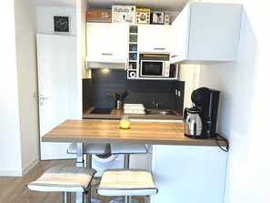 Eine kleine Küche mit Mikrowelle, Kaffeemaschine und einem Spülbecken. Zwei Barhocker und ein Stuhl sind vorhanden.