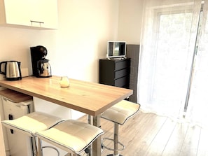 Eine kleine Küchenzeile mit Kaffeemaschine, Wasserkocher und einem Fernseher auf einem Schrank. Zwei Barhocker und ein Holztisch sind vorhanden.