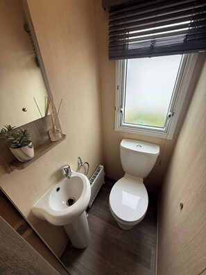 Casa de banho compacta com sanita, bidé e lavatório. Tem uma janela com persianas, um radiador e uma pequena estante com plantas.