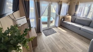 A modern living room with a sofa, a flat-screen TV, a heater, and a sliding glass door leading to a pool. - Fantasy Island H12 Caravan With Hot Tub (Skegness)