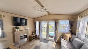A cozy living room with a flat-screen TV, a fireplace, a sofa, and a view of a hot tub outside. - Fantasy Island H12 Caravan With Hot Tub (Skegness)