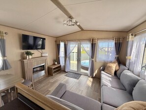 A cozy living room with a flat-screen TV, a fireplace, a sofa, and a view of a hot tub outside. - Fantasy Island H12 Caravan With Hot Tub (Skegness)