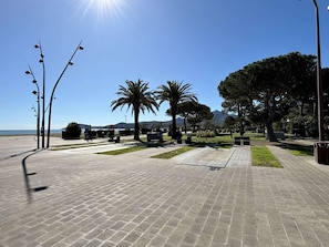 Ein gepflasterter Weg mit Bänken, Palmen und freiem Blick auf das Meer und die dahinterliegenden Berge.