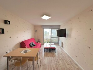 A compact living space with a dining table, a red sofa, a TV mounted on the wall, and a view through a large window. - Cozy apartment near the sea - 4 guests (Courseulles-sur-Mer)