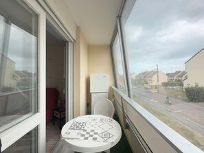 A balcony with a table, a chair, a refrigerator, and a view of a street with houses and a motorcyclist. - Cozy apartment near the sea - 4 guests (Courseulles-sur-Mer)