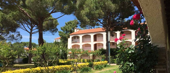 A multi-story building with a red-tiled roof, surrounded by lush greenery and yellow flowering plants.