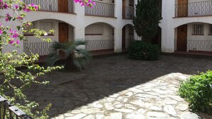 Un cortile con pavimento in pietra, pareti bianche, balconi con ringhiere in ferro battuto e piante fiorite.