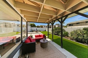 Eine überdachte Terrasse im Freien mit einer Couch und einem runden Tisch, einem Rasen und Blick auf benachbarte Häuser.