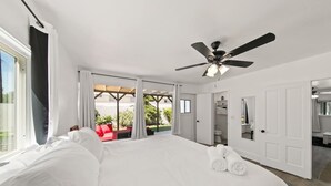 Un grand lit avec du linge de lit blanc, un ventilateur de plafond et une vue sur une terrasse avec une chaise rouge et des plantes.