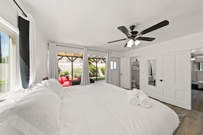 Ein großes Bett mit weißen Leinen, ein Deckenventilator und Blick auf eine Terrasse mit einem roten Stuhl und Pflanzen.