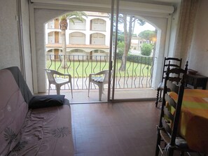 A room with a sofa, a dining table, and chairs, a balcony with a view of a building and greenery. - Garden-side apartment - 4 guests (Argelès-sur-Mer)