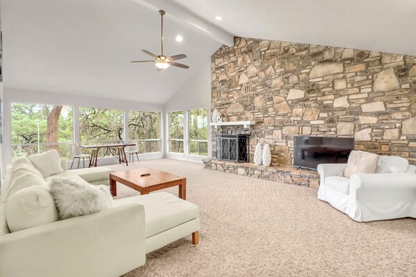 A spacious living room with a stone fireplace, white furniture, and large windows offering a view of trees. - 16 Private Acres Near Wimberley Square | Hot Tub & Room for Groups (Wimberley)
