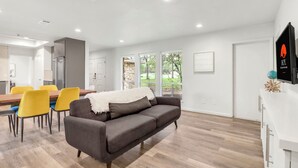 Una sala moderna con un sofá gris, sillas amarillas, una mesa de comedor de madera y una cocina al fondo.