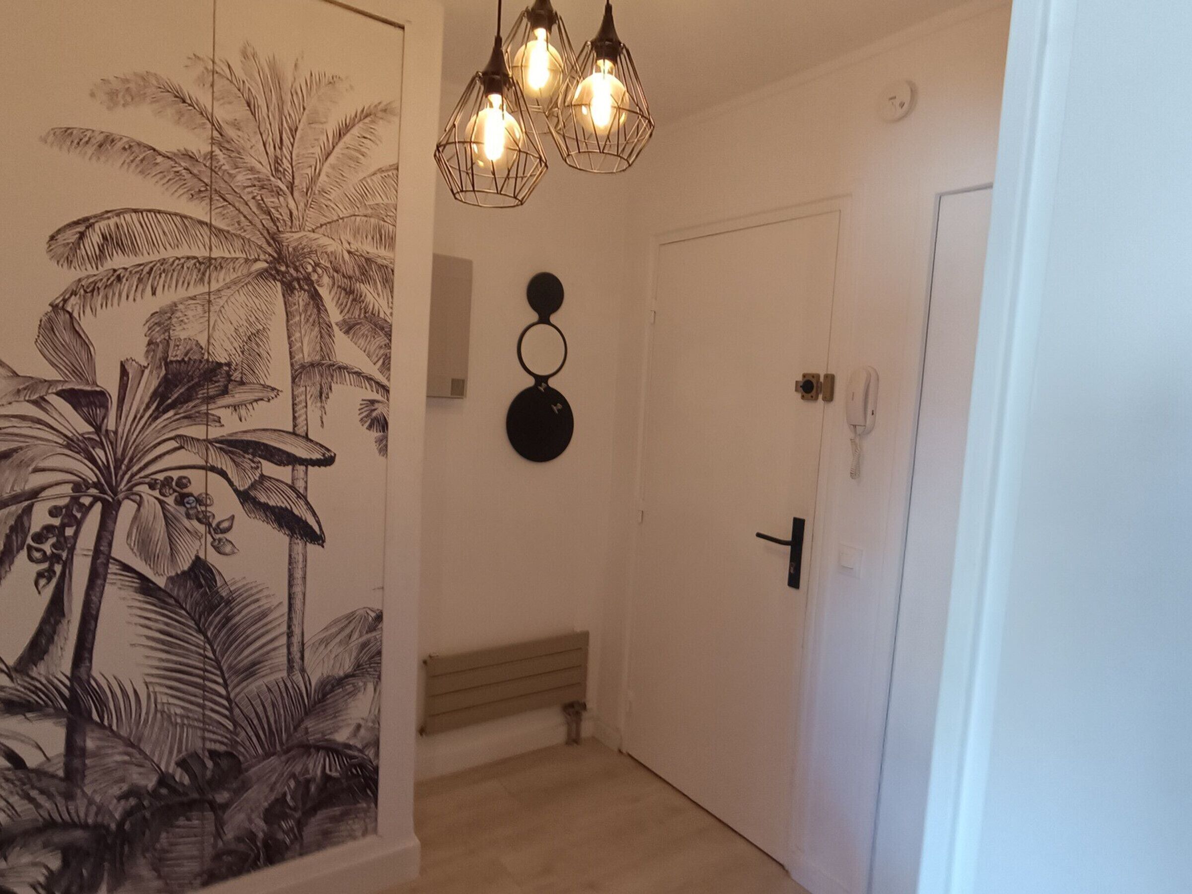 Um corredor com uma porta, um luminário fixado na parede e uma arte na parede com palmeiras.