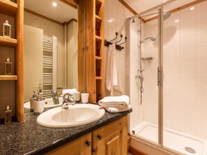 Une salle de bain avec un lavabo surmonté d’un plan de travail en granit, des meubles en bois, une douche à parois de verre et un miroir avec des étagères.