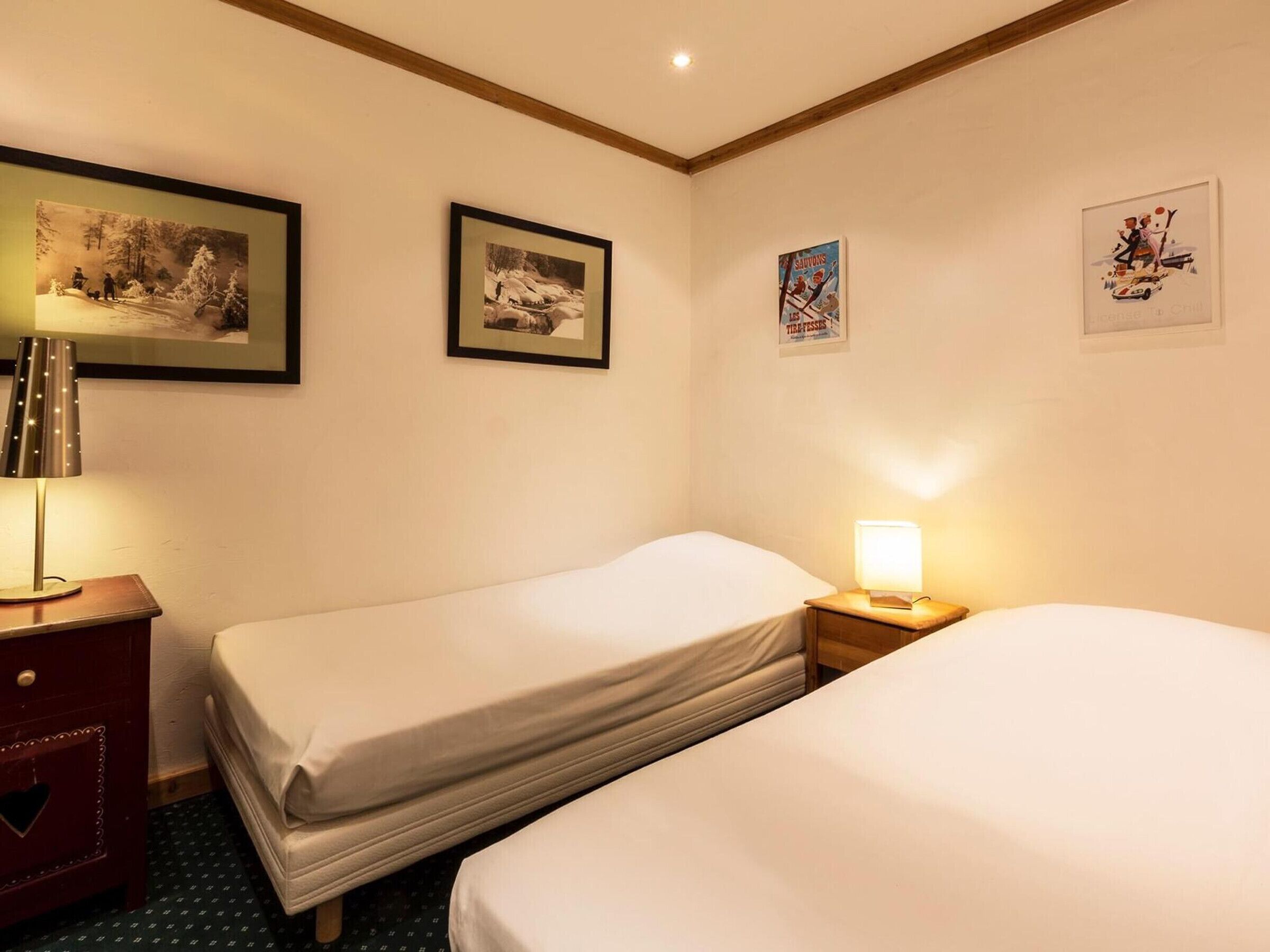 Quarto de hotel com duas camas, uma mesa de cabeceira com abajur e quadros nas paredes.