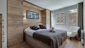 Une chambre moderne avec un mur en bois, un lit recouvert d’une couverture marron, une chaise blanche et une fenêtre donnant sur des immeubles.